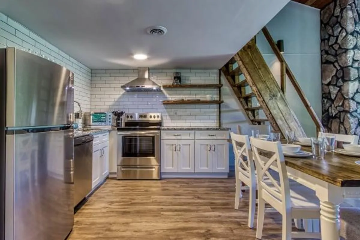 A bright, open kitchen featuring stainless steel appliances, granite countertops, and open shelving. The wooden dining table sits nearby, creating a seamless flow between the cooking and dining spaces. Warm lighting and natural wood accents make the space feel both functional and welcoming.
