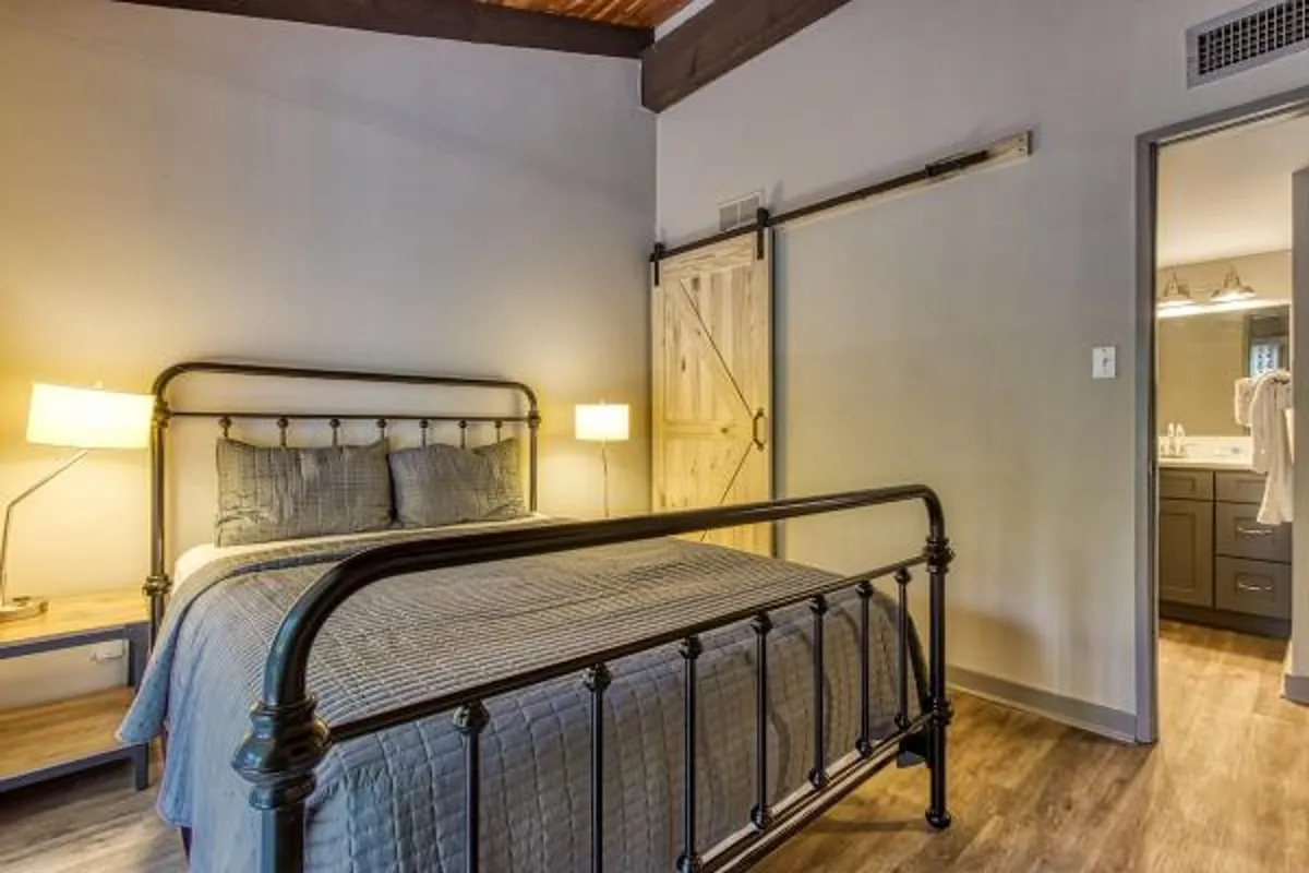 A serene bedroom with a metal bed frame, decorative pillows, a bedside lamp, and a rustic sliding barn door. Sunlight filters in, highlighting the inviting textures and intimate ambiance.