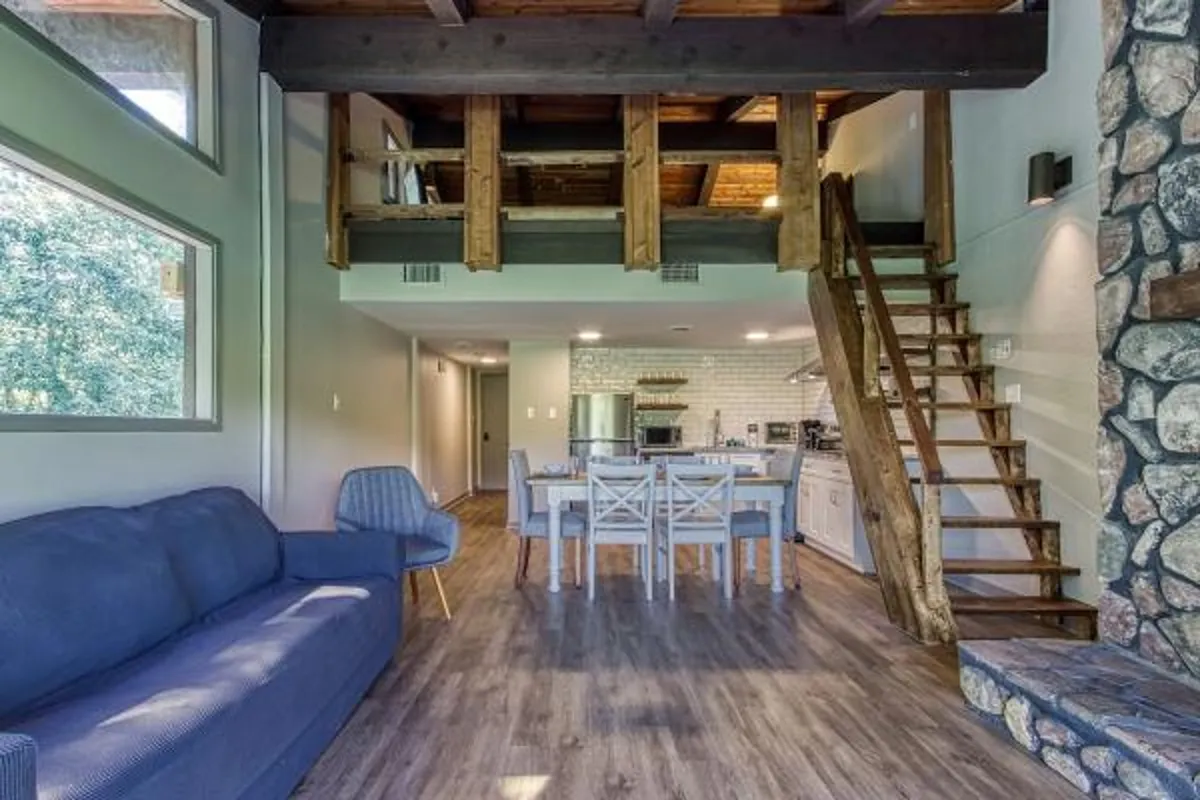 A cozy living room featuring a comfortable sofa, a stone fireplace, and large windows that let in natural light. The space exudes a rustic yet modern cabin vibe, with wood accents and an inviting layout perfect for family gatherings or peaceful relaxation.