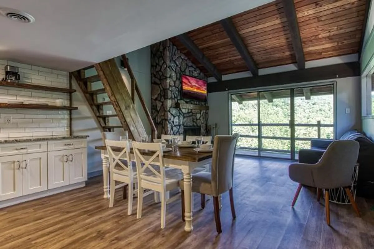 A cozy dining area set near a stone fireplace. The room is filled with natural light from large windows, and a wooden dining table is ready for meals or gatherings. The open staircase and sightlines into adjacent spaces reinforce the sense of connection.