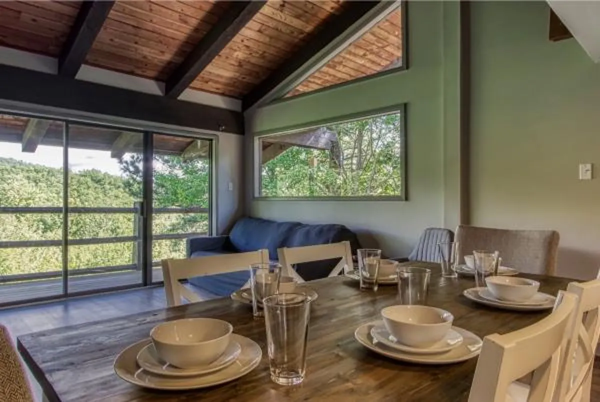 A dining table elegantly set with plates and glasses, positioned by expansive windows that offer lush green views. A comfortable sofa in the corner provides additional seating, and the overall atmosphere is inviting and social.