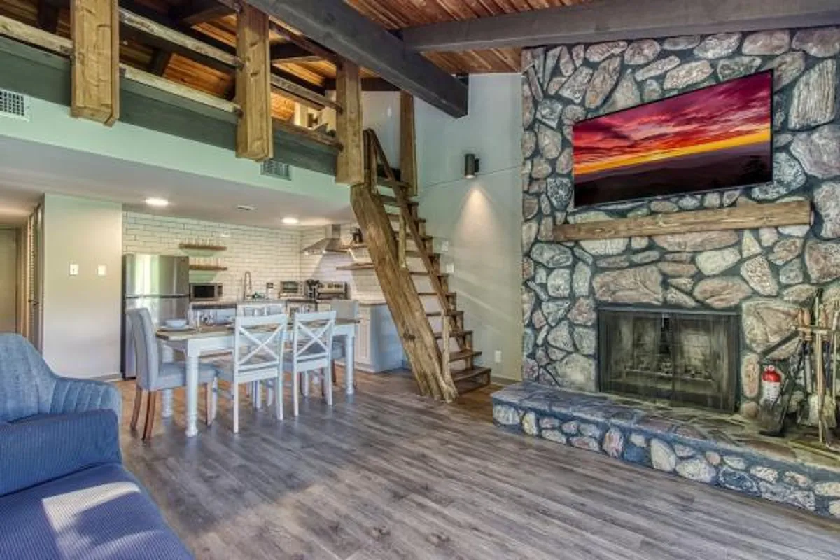 A warm living space with a stone fireplace, wooden beams, dining table, and modern touches like a wall-mounted TV. The layout emphasizes communal areas perfect for meals, movie nights, or relaxing by the fire, blending rustic details with contemporary comfort.