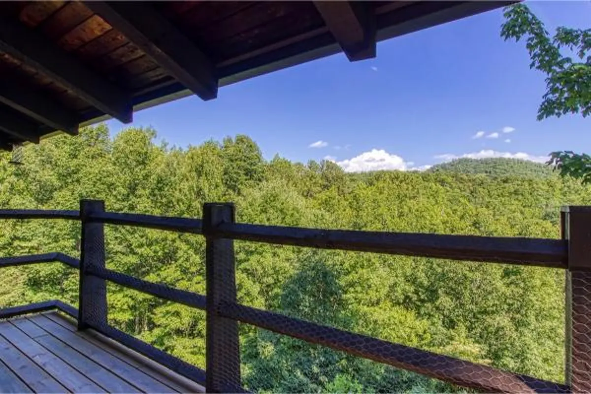 A serene perspective from a wooden deck railing overlooking lush greenery and distant rolling hills, with clear skies creating a welcoming, open feel. The vantage point suggests a relaxing retreat connected to nature.