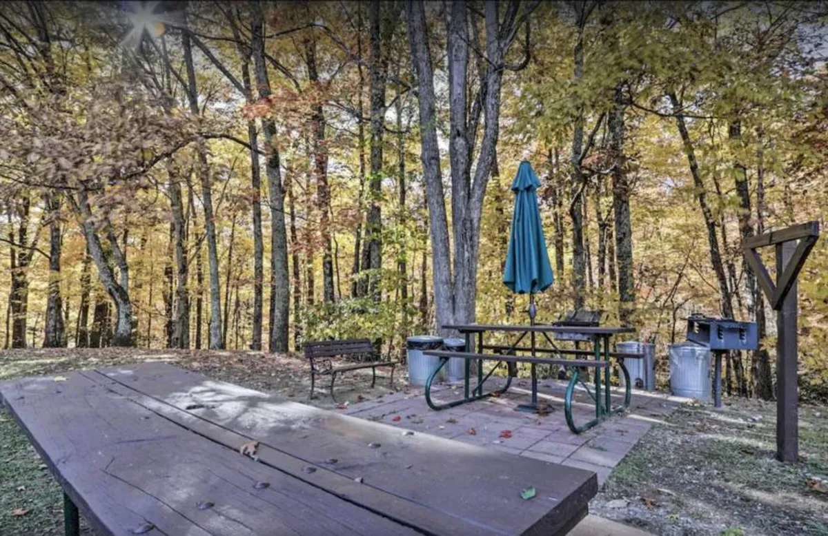 An inviting outdoor scene featuring a wooden picnic table under a green umbrella, surrounded by trees and subtle fall colors. The area is set up for casual dining or a morning coffee, evoking a cozy, intimate atmosphere for couples.